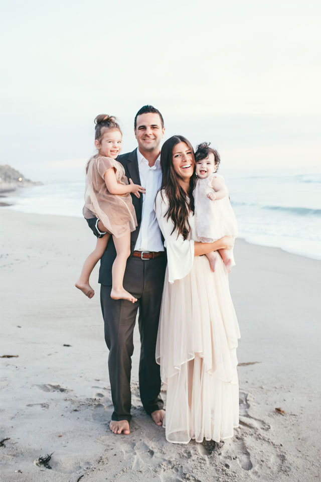 Smiling man and woman both holding children in formal attire posing for photo barefoot, outdoors on the beach with ocean in background