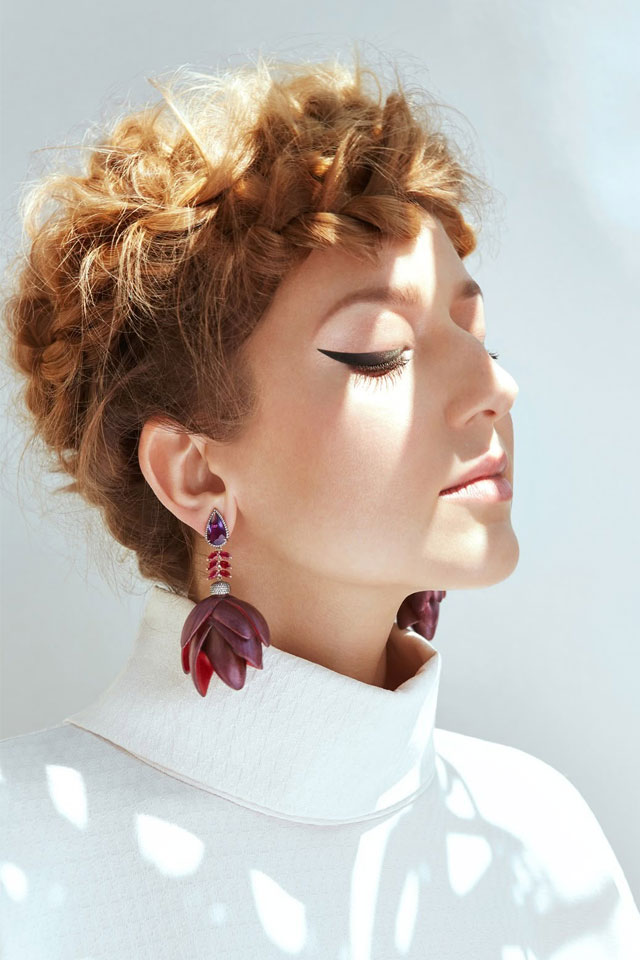 Woman wearing white top with short, blonde hair and head tilted upward towards rays of sunlight with eyes closed, wearing purple and red chunky botanical earrings