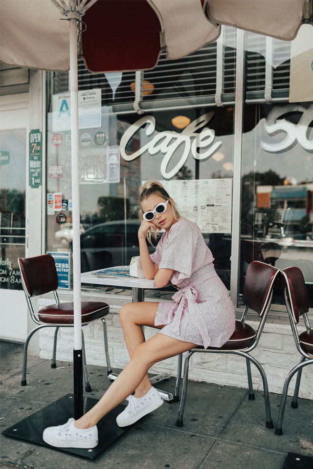 young woman with blonde hair, white-rimmed sunglasses wearing a pink and white pinstriped sundress and sitting resting her head on her hand at an outdoor table at a cafe