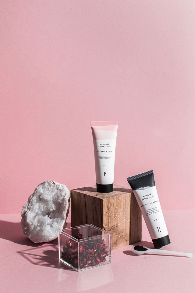 Stylised photo of 2 skincare product tubes against pink backdrop on wooden block with quartz crystal geode, botanics and a spoon