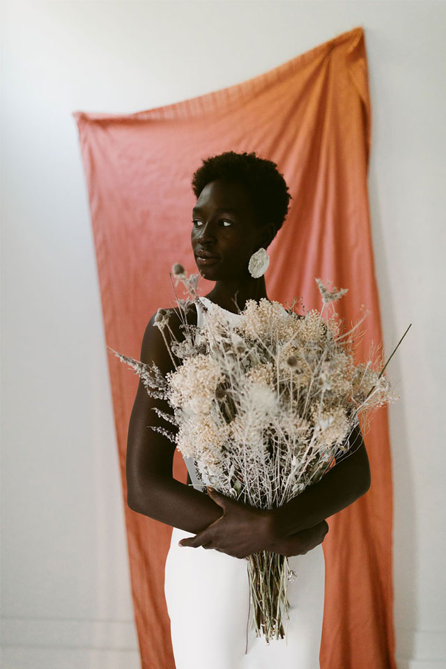 portrait view of a woman looking off to the side and holding a large bunch of dried flowers in front of an orange backdrop