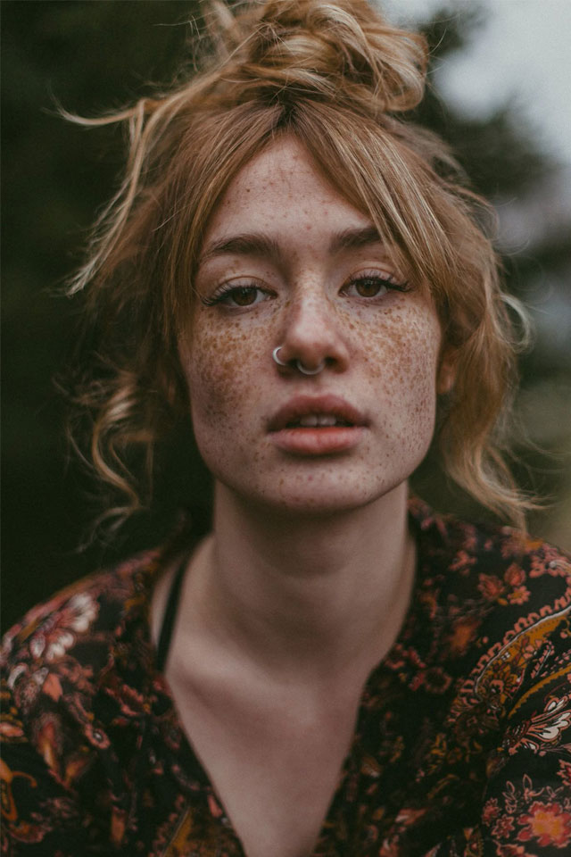 close-up image of young woman with hair in a messy bun, freckles, nostril and septum ring against a autumnal moody blurred background