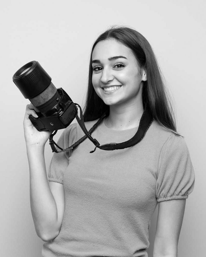Portrait photo of young woman with long hair in a pink top, smiling and holding a camera