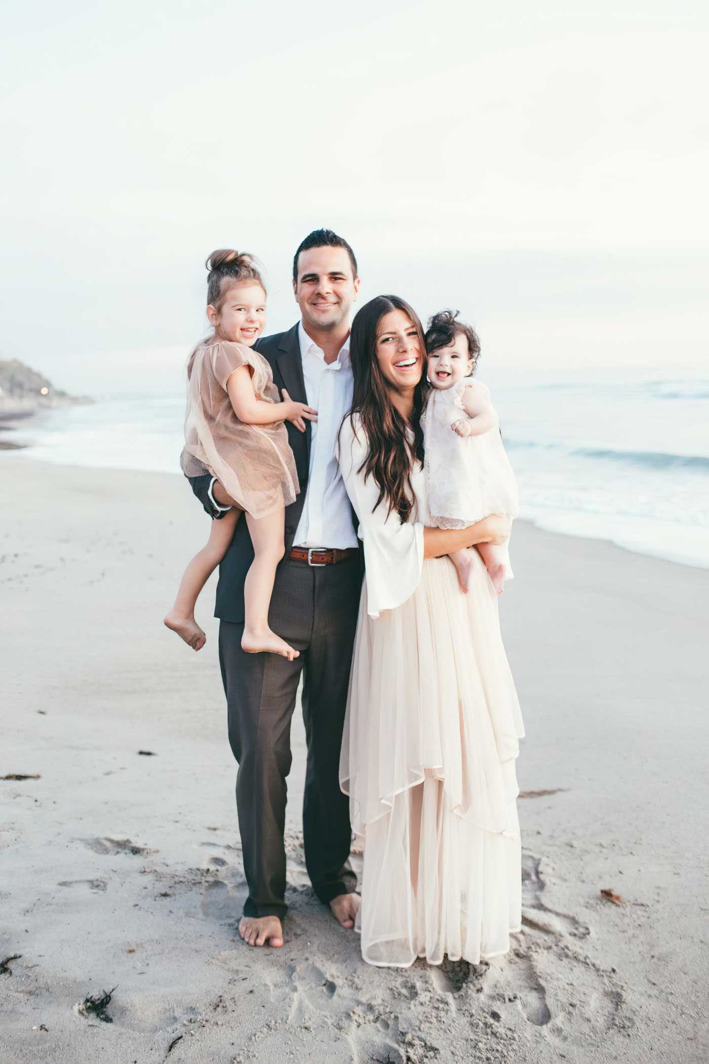 Smiling man and woman both holding children in formal attire posing for photo barefoot, outdoors on the beach with ocean in background