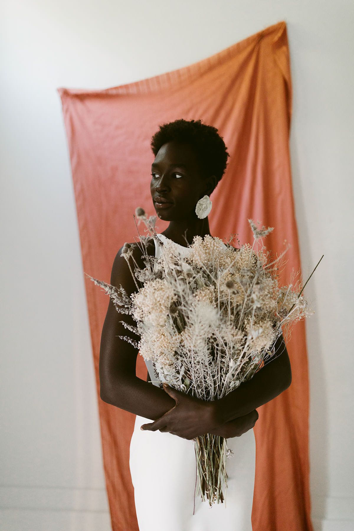 portrait view of a woman looking off to the side and holding a large bunch of dried flowers in front of an orange backdrop