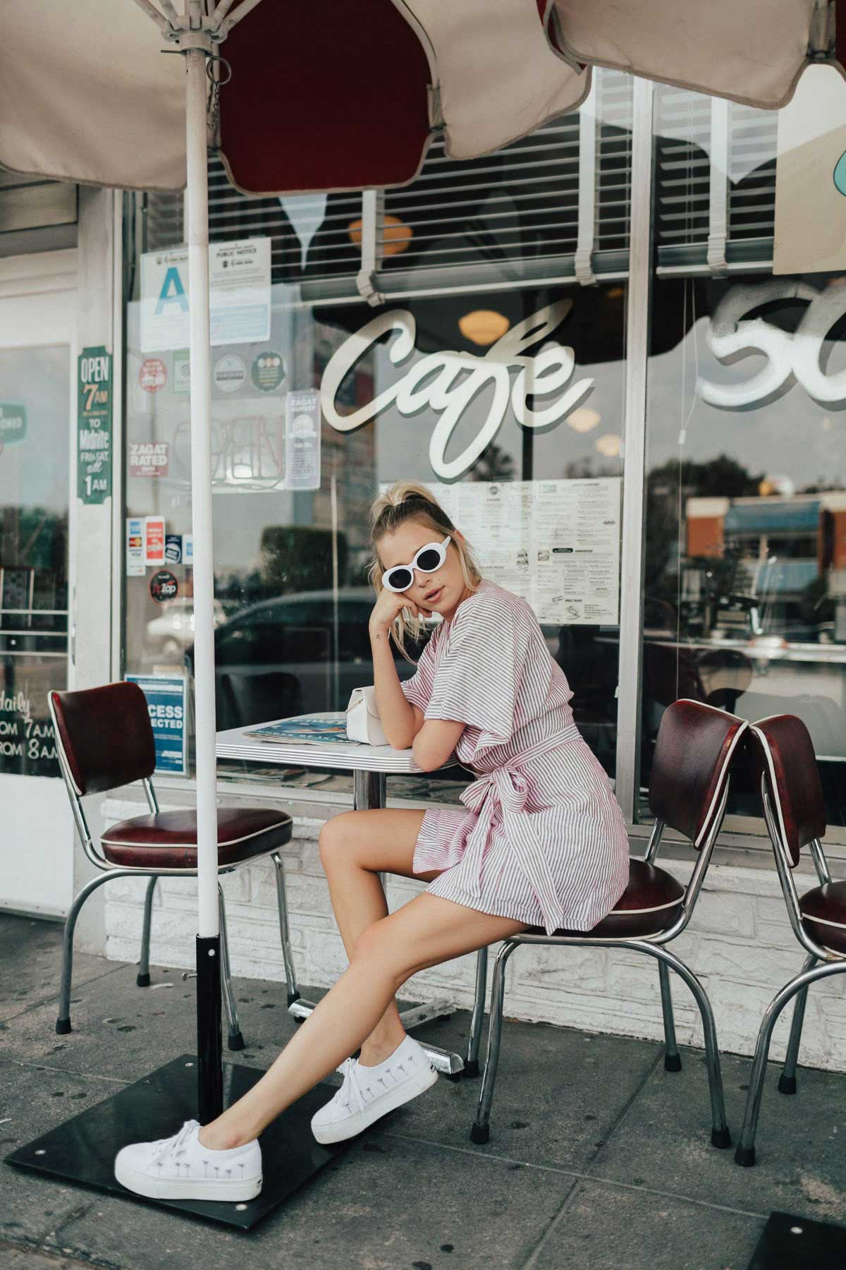 young woman with blonde hair, white-rimmed sunglasses wearing a pink and white pinstriped sundress and sitting resting her head on her hand at an outdoor table at a cafe