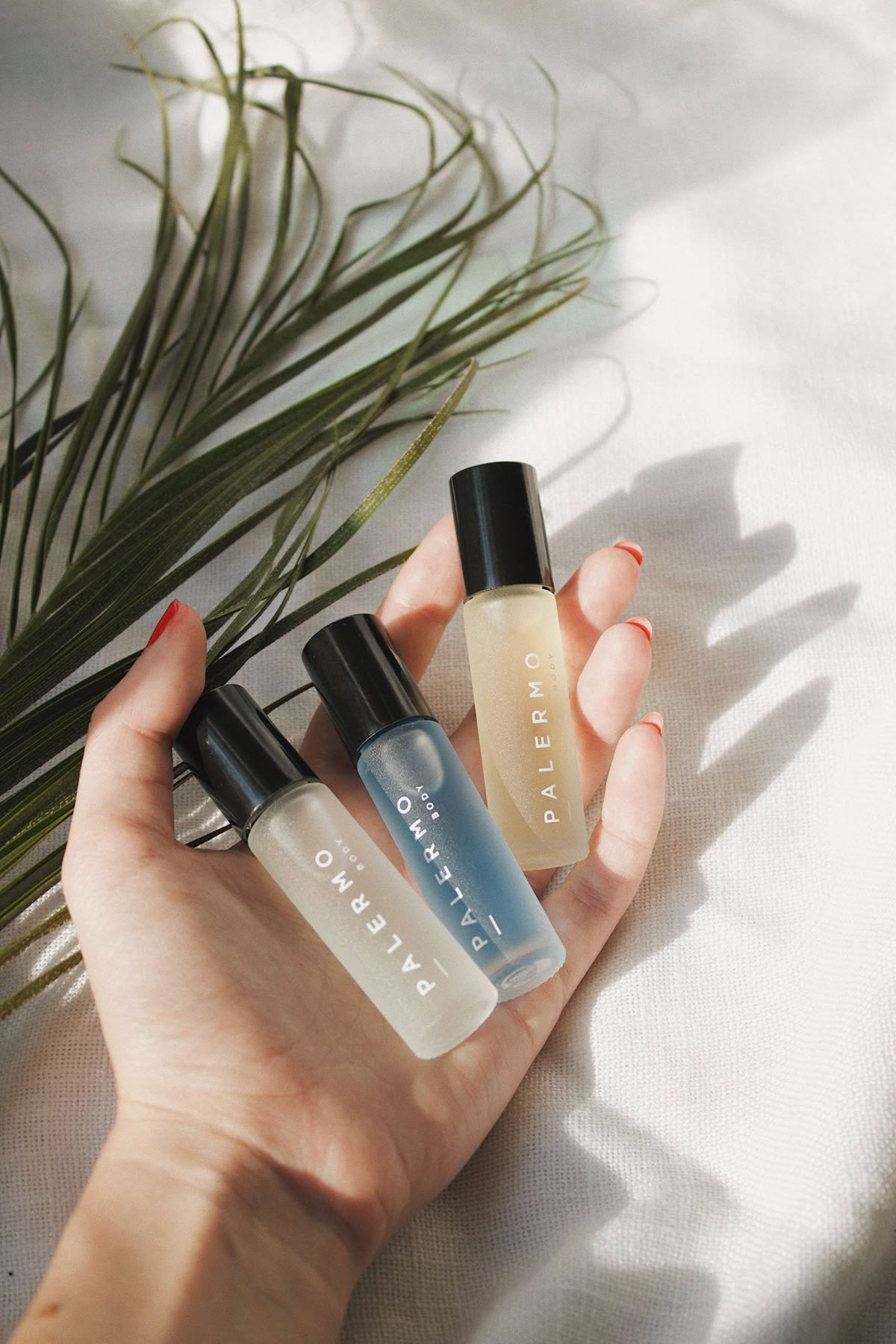 Woman's hand with coral nail colour lays on white fabric backdrop and a fern holding 3 small travel bottles of Palermo body products with black lids