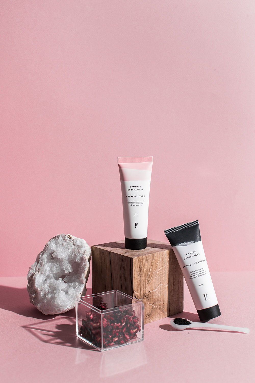 Stylised photo of 2 skincare product tubes against pink backdrop on wooden block with quartz crystal geode, botanics and a spoon