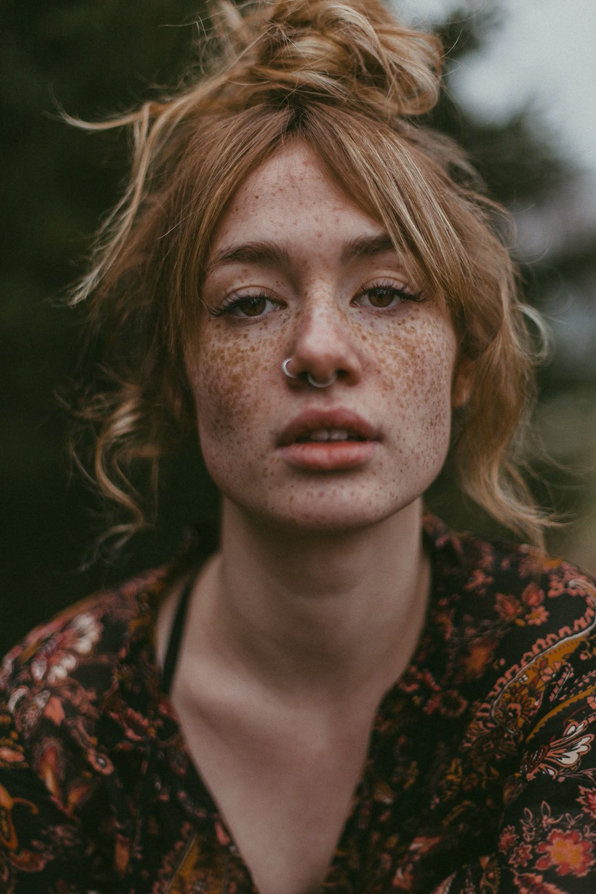 close-up image of young woman with hair in a messy bun, freckles, nostril and septum ring against a autumnal moody blurred background